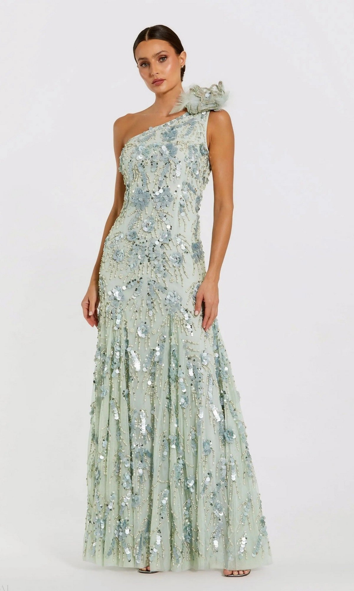 A woman stands wearing the Mac Duggal 94151 long formal dress in sage, a floor-length, one-shoulder designer gown embellished with sequin beaded details and floral accents, posing against a plain white background.